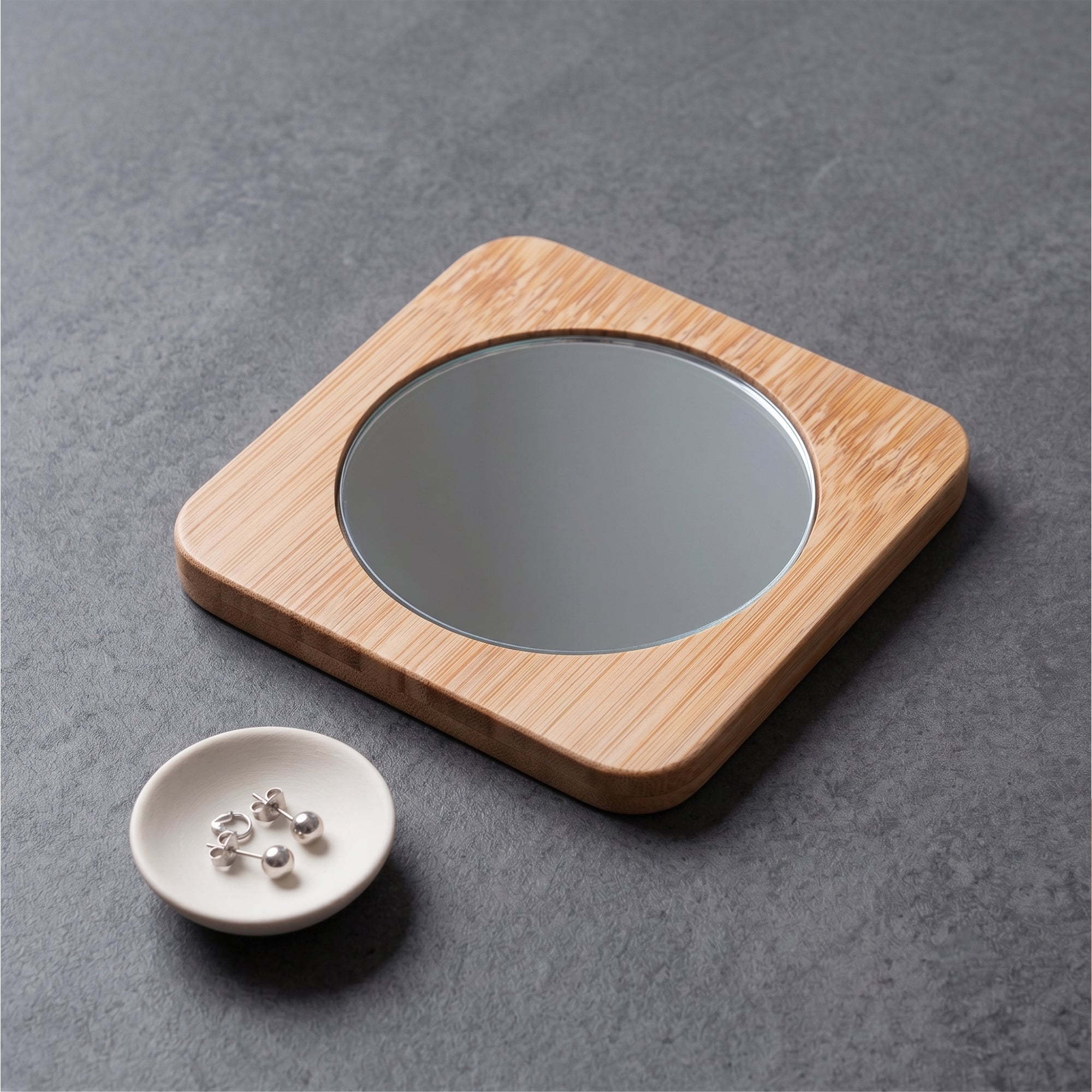Espejo de bambú natural apoyado sobre una superficie gris junto a un pequeño plato con aros